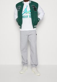 Chaqueta varsity verde y blanca con letras en blanco y negro, camiseta gráfica blanca, pantalones de chándal grises y zapatillas color crema.