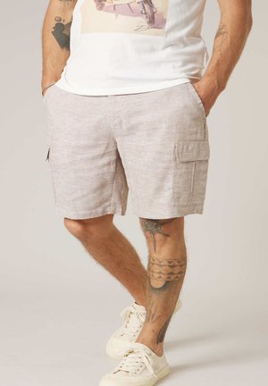 Homme portant un short cargo beige, un t-shirt blanc graphique, des baskets blanches, avec des tatouages sur les deux bras et les jambes, debout les mains dans les poches.