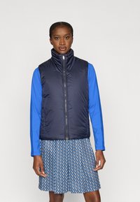 Colete azul-marinho sem mangas com gola alta, fecho de correr e tecido texturizado, combinado com uma camisa azul de manga comprida e uma saia estampada.