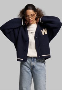Marineblauer gerippter Cardigan mit weißen Akzenten und großem 'W'-Emblem, ausgestattet mit Knopfverschluss und lockerer Passform, kombiniert mit hellen Jeans.