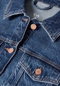 Veste en denim bleu avec boutons en cuivre, col visible, poche poitrine et surpiqûres marron.