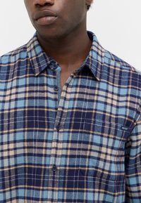 Blauw en marine geruit shirt met beige accenten, met een knoopsluiting, kraag en klein logo op de borst. Zachte textuur.