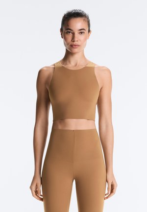 Žena nosiaca bezrukávový, krátky telový športový top a telové legíny s vysokým pásom na jednoliatom svetlom pozadí.