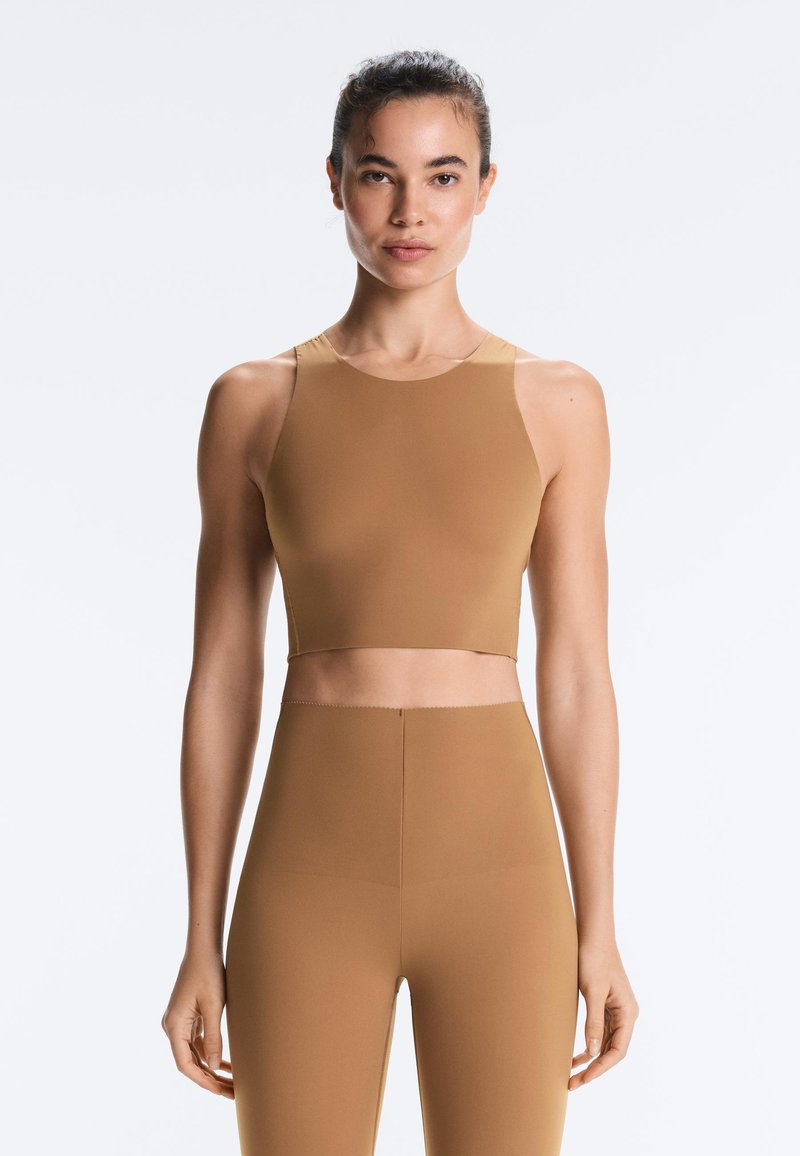 Femme portant un haut de sport court sans manches beige et des leggings taille haute beige sur un fond clair uni.