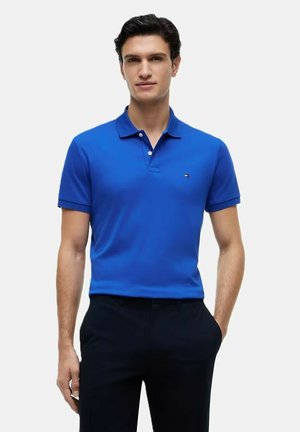 Giovane uomo che indossa una polo a maniche corte blu brillante e pantaloni neri, in piedi con una mano in tasca contro uno sfondo semplice.