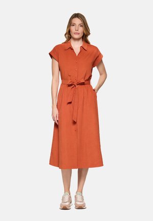 Mujer de pie con un vestido midi color óxido con botones, cinturón anudado a la cintura, mangas cortas y zapatillas beige, sobre un fondo claro y liso.