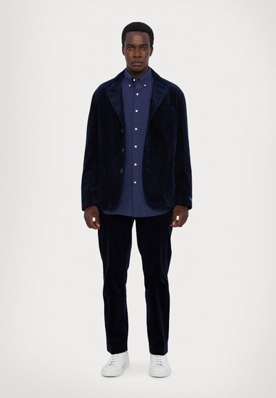 Marineblå kordfløyelsblazer og bukser, kombinert med en blå prikkete skjorte. Hvite sneakers fullfører antrekket. Jevn tekstur gjennomgående.