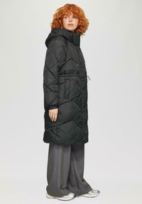 Zwarte gewatteerde pufferjas met een capuchon, aangesloten taille en matte afwerking; heeft diagonale stiksels en zijzakken. Gepaard met grijze broeken.