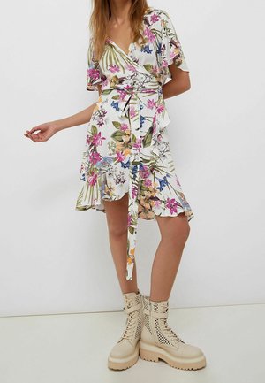 Femme portant une robe portefeuille blanche à fleurs à manches courtes avec une ceinture nouée à la taille et des bottines beiges à lacets avec des détails ajourés.