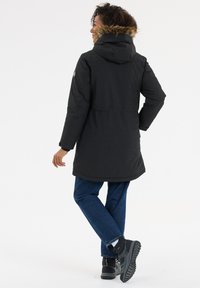 Zwarte parka-jack met een bont-achtig gevoerde capuchon, met een relaxed fit, lange mouwen en een trekkoord in de taille, gedragen over blauwe spijkerbroeken.
