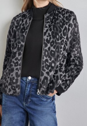 Kobieta ubrana w szaro-czarną kurtkę na suwak w panterkę, na czarnym swetrze i niebieskich dżinsach, z jedną ręką w kieszeni.