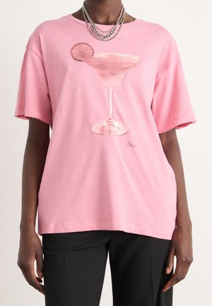 Person iført en pink T-shirt med et print af et pink cocktailglas med en citronbåd, kombineret med lagdelte sølvkæder og sorte bukser.