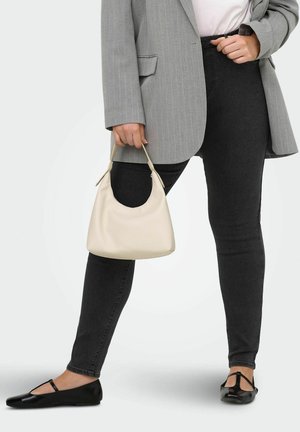 Kvinde i sorte skinny jeans, grå blazer med tynde striber og sorte flade sko, der holder en lille beige håndtaske mod en lys baggrund.