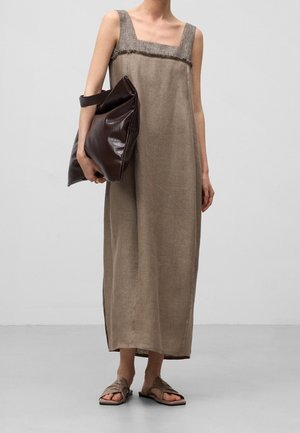 Frau trägt ärmelloses beiges Maxikleid und hält eine große dunkelbraune Ledertasche, mit geflochtenen Sandalen auf grauem Boden.