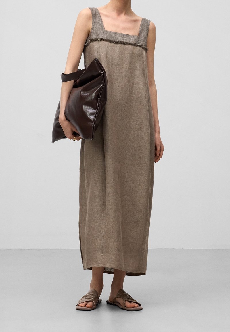 Femme portant une robe maxi beige sans manches, tenant une grande pochette en cuir marron foncé, avec des sandales tressées sur un sol gris.