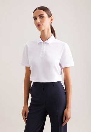 Kvinde iført en hvid kortærmet poloshirt stoppet ned i mørkeblå bukser med høj talje, stående mod en ensfarvet baggrund.