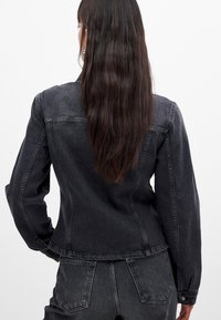 Veste en denim noire à la coupe ajustée, manches longues et col classique. Le tissu présente une finition légèrement texturée et des coutures visibles.
