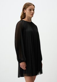 Zwarte geplooide jurk met lange, transparante mouwen en een ronde halslijn, met een vloeiende silhouet en zachte textuur.