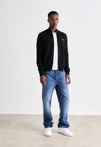 Man standing against white background wearing a black zip-up jacket, white t-shirt, blue jeans, and white sneakers.