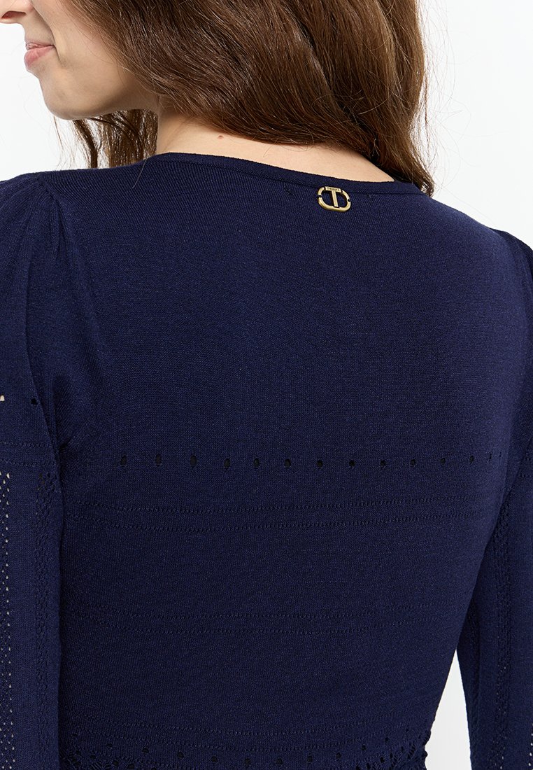 Pull en tricot bleu marine avec des détails ajourés délicats aux manches et un accent logo doré au dos du col.