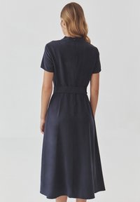 Marineblauwe midi-jurk met korte mouwen, een getailleerde taille en een uitlopende rok. Heeft een verborgen ritssluiting aan de achterkant en een gladde textuur.