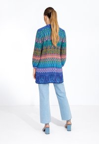 Multicolored long blouse with a geometric pattern, featuring shades of blue, green, and pink. Worn with light blue wide-leg jeans and heels.