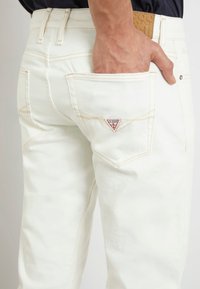 Vaqueros de denim blanco con costuras naranjas, un logotipo triangular rojo en el bolsillo trasero derecho y un cinturón texturizado color beige visible encima de la cintura.