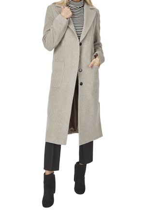 Kvinde iført en lang beige frakke med knapper, sorte cropped bukser, sorte ankelstøvler og en sort-hvid stribet rullekrave.