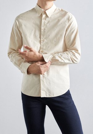 Mężczyzna poprawia mankiet beżowej koszuli na długi rękaw z guzikami, ubrany w ciemne spodnie, stoi na tle jasnego, jednolitego tła.