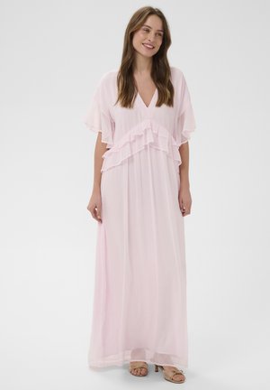 Jeune femme souriante, portant une robe maxi fluide rose clair avec des détails à volants et des talons ouverts beiges, debout devant un fond uni.