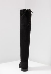 Black suede over-the-knee boot with a flat sole; features a rounded toe and adjustable tie at the top for fit. Smooth texture and sleek design.