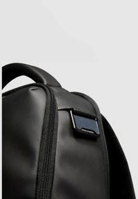 Schwarzer Rucksack mit glatter, matter Oberfläche, einem Reißverschlussfach und einem auffälligen Metallelement am Träger mit Branding-Detail.