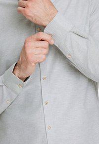 Camicia grigia chiaro con una sottile texture, dotata di bottoni beige e colletto su misura. È visibile un polsino che viene regolato.