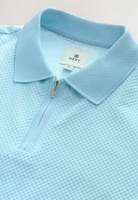 Tricou polo de culoare albastră deschisă cu un model texturat în formă de diamant, având un guler cu fermoar și etichetă cu inscripția „NEXT”. Material neted, cu plasă respirabilă.