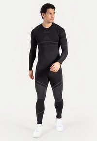 Schwarzes langärmliges Sport-Oberteil und enganliegende Leggings mit strukturierten Akzenten und kontrastierenden Nähten. Kombiniert mit weißen Sportschuhen.