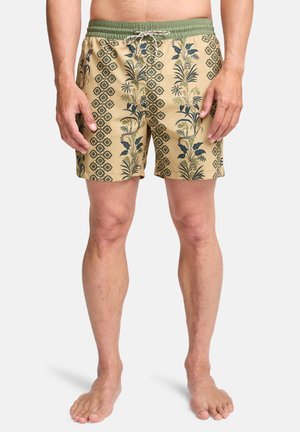 Man die beige zwembroek draagt met groene bloemen- en geometrische patronen, barefoot staand tegen een effen witte achtergrond.