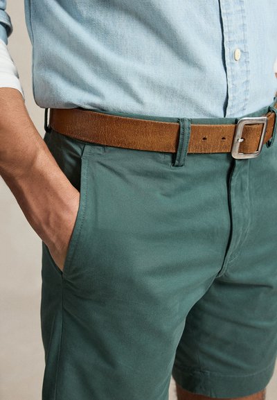 Bărbat care poartă pantaloni scurți turcoaz cu buzunare în față, o curea maro din piele și o cămașă bleu cu nasturi băgată în pantaloni, cu mâna în buzunar.