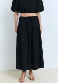 Jupe maxi noire en tissu léger texturé, avec une ceinture à la taille et un design en étages. Assortie avec un haut court coordonné et des sandales.