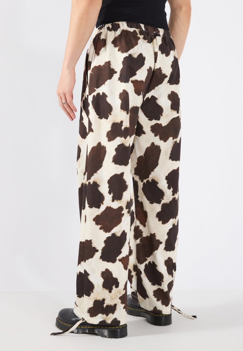 Pantalon à jambes larges avec un imprimé vache marron et noir, porté avec des bottes noires et un haut noir, vu de dos à mi-mollet.