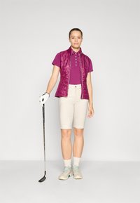 Kurzärmliges lila Poloshirt, hellbeige Shorts, bordeauxfarbener gesteppter Weste, weißer Golfhandschuh, Golfschläger, hellfarbene Golfschuhe mit türkisfarbenen Akzenten.