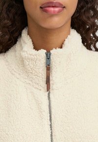 Helebeež fleece-jakk kõrge kraega, millel on hõbedane tõmblukk ja pruun nahast tõmblukk. Pehme, tekstureeritud pind, millel on nähtavad õmblused.