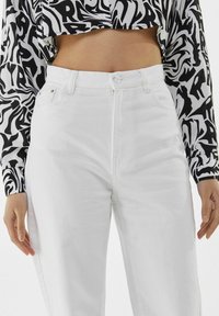Jeans blancos de cintura alta con una ligera textura, que cuentan con passadores para cinturón, bolsillos frontales y un cierre de botón, combinados con una blusa estampada en blanco y negro.