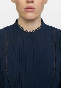 Tumes sinine pluus vertikaalsete voldikutega, nö�öb закрытие ja tekstureeritud pitsist kaelus. Omab külgpitsist sisendeid ja sileda kangaste viimistlust.