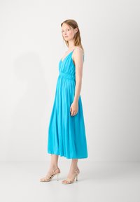 A sleeveless, V-neck, long dress in bright turquoise fabric, featuring a gathered waistline and flowing skirt. Paired with light beige strappy heels.