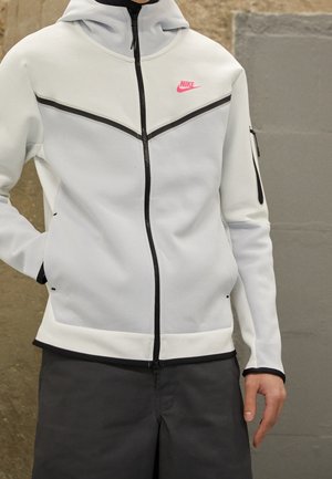 Osoba nosząca białą bluzę z kapturem Nike na zamek, z czarnymi wykończeniami i różowym logo, w połączeniu z ciemnymi spodniami, stojąca na tle teksturowanej ściany.