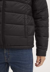 Jachetă puffer neagră cu închidere prin fermoar, design matlasat și manșete elastice. Purtați-o cu blugi din denim albastru, evidențiind un croi relaxat.