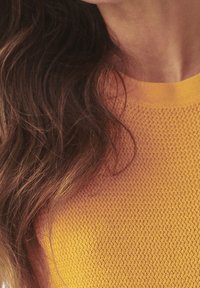 Langt brunt hår over skulderen, iført en tekstureret gul sweater med rund halsudskæring, delvis udsigt til nakke og hage.