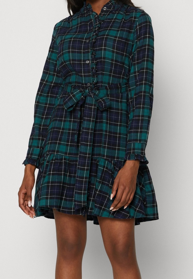 Femme portant une robe à manches longues à carreaux verts et bleu marine avec des boutons, des détails volantés et une ceinture nouée à la taille, debout devant un fond blanc.