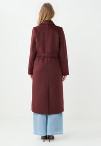 Cappotto in misto lana bordeaux con vita cintata, maniche lunghe e silhouette dritta, abbinato a pantaloni in denim a gamba larga.