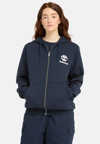 Sudadera con cremallera en azul marino hecha de tela suave, con un logo blanco de Timberland en el lado izquierdo del pecho y bolsillos tipo canguro.